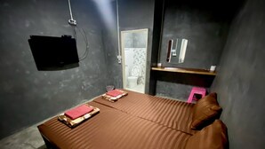 A room with a bed, a small table, a mirror, and a wall-mounted TV. - Ploynam Homestay (Amphawa)
