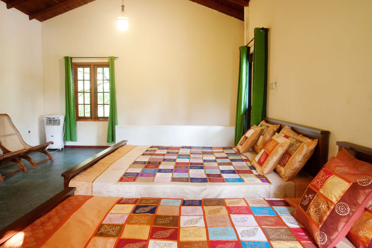 Quarto com uma cama grande, uma cadeira de madeira, janela com cortinas verdes e ventilador de teto.