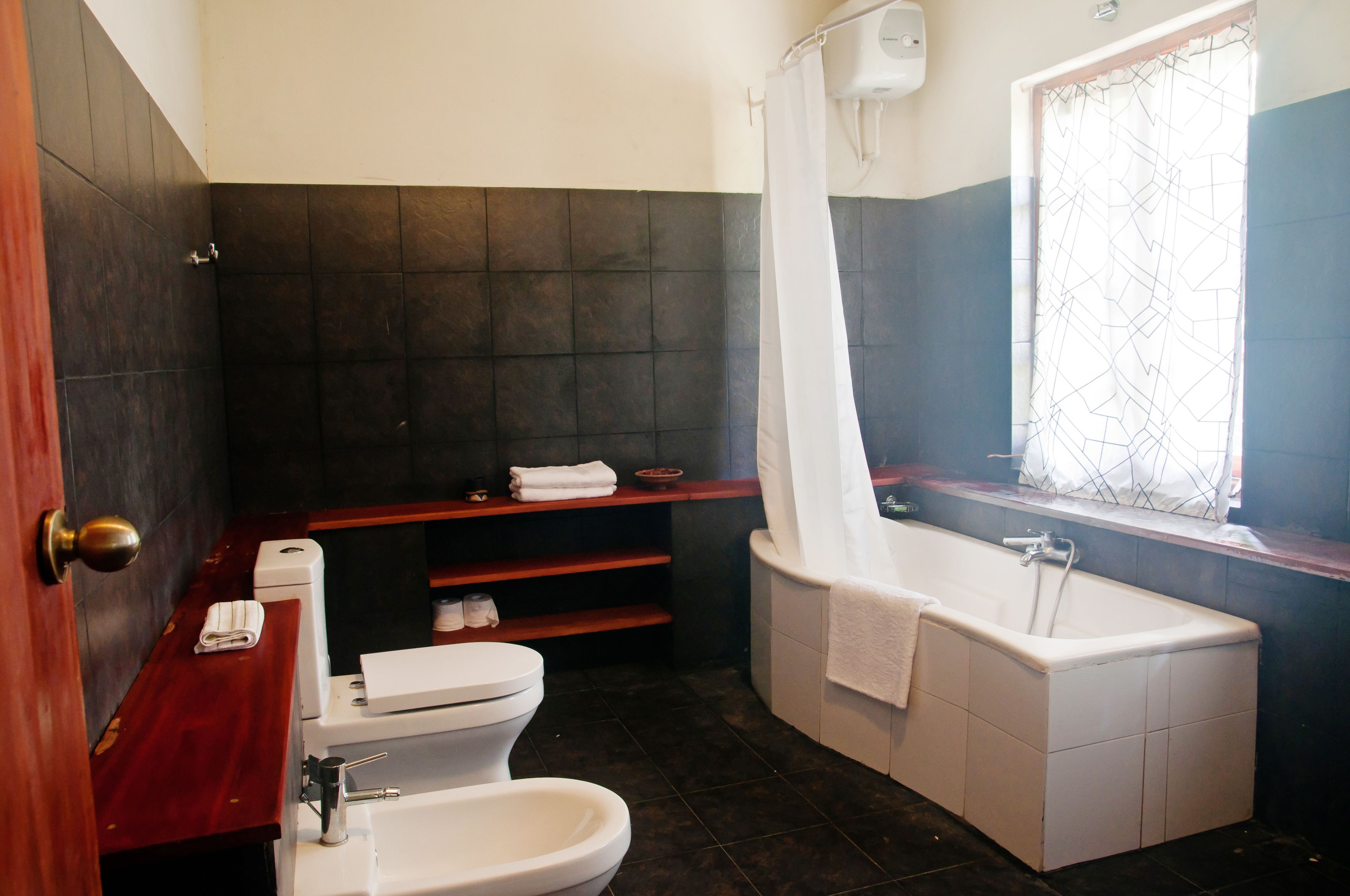 Une salle de bain avec une baignoire blanche, des toilettes et un lavabo. Il y a une fenêtre avec un rideau et une étagère avec des serviettes.