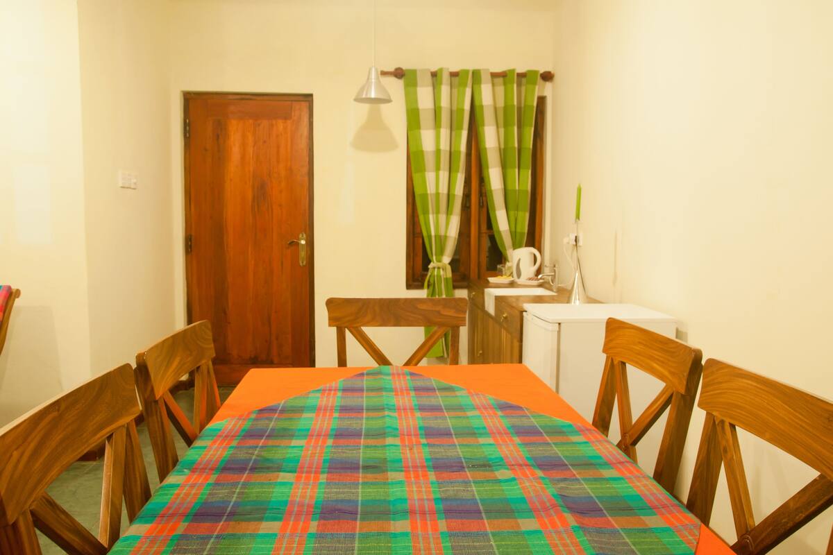 Uma área de refeições com uma mesa posta para quatro pessoas, cadeiras de madeira, toalha xadrez, uma porta de madeira, uma janela com cortinas e um pequeno armário branco.