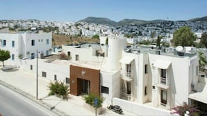 A modern building with a flat roof, a central entrance, and a view of a residential area with white houses and a mountain in the background. - New Bodrum Hotel (Bodrum)