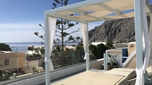 A balcony with a white canopy, beige cushions, and a view of the sea and mountains.