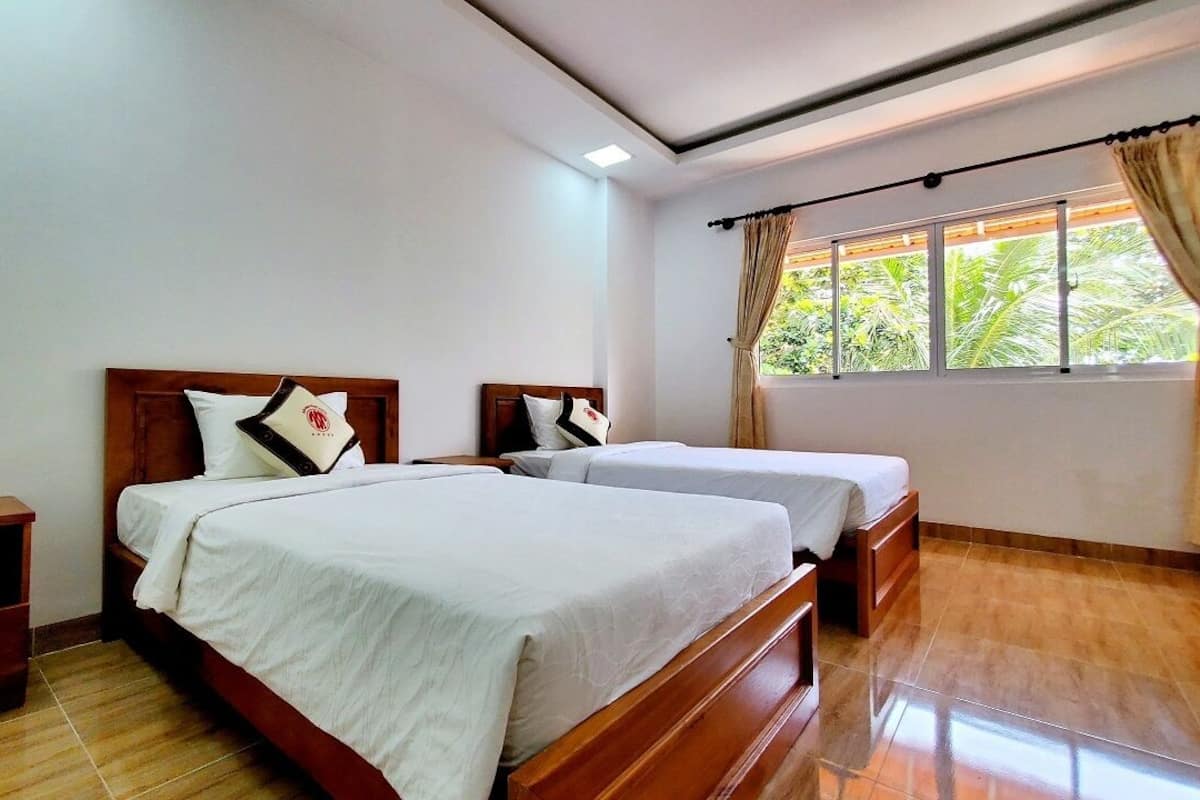 A hotel room with two beds, wooden headboards, white bedding, a nightstand, and a window with curtains.