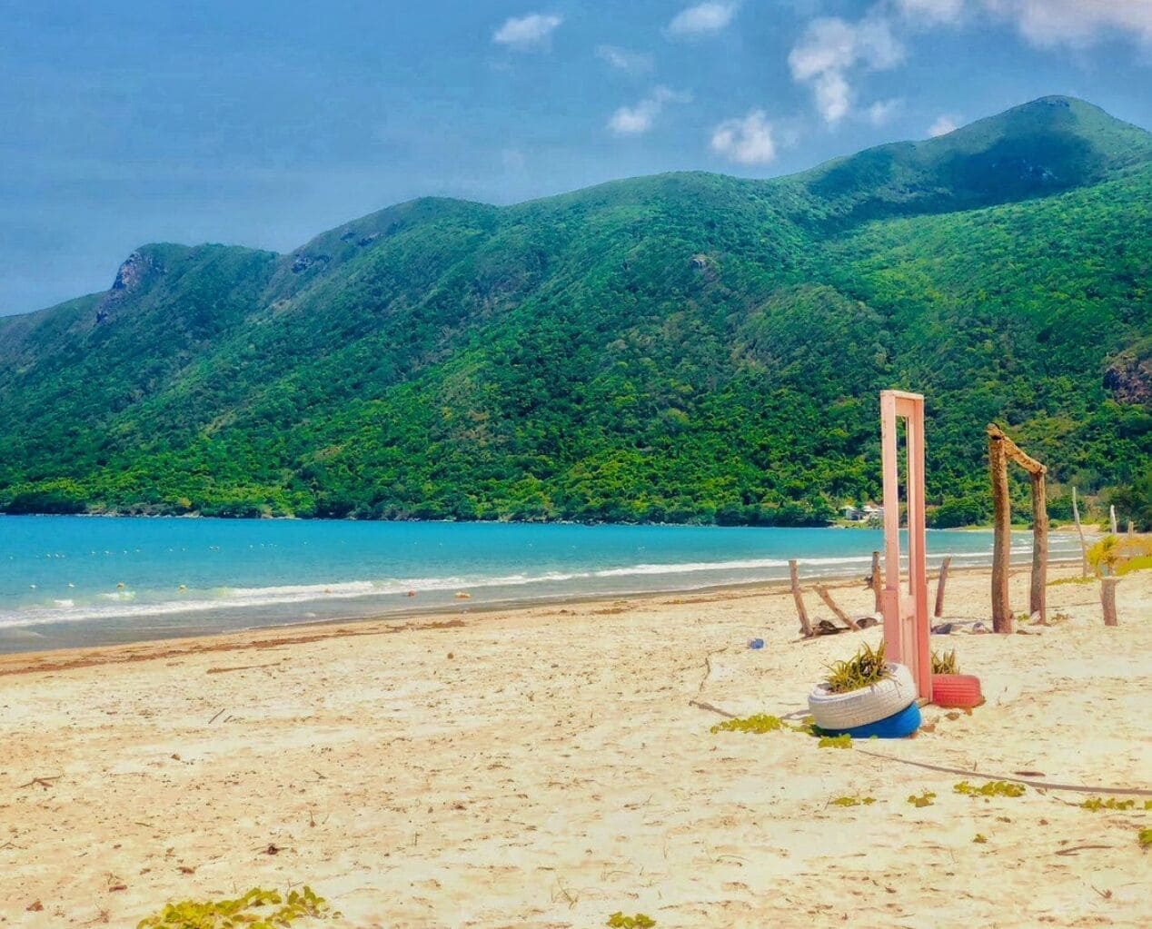 Una playa con orilla arenosa, mar azul cristalino y colinas verdes al fondo.