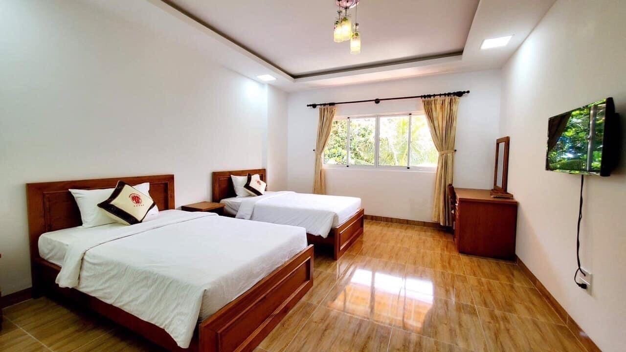 A hotel room with two single beds, a TV mounted on the wall, a wooden nightstand, and a window with curtains.