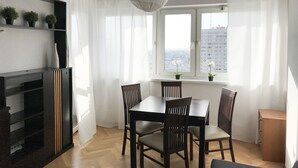 Un'area pranzo moderna con un tavolo e sedie in legno, una lampada a soffitto bianca e vista sulla città attraverso una finestra con tende leggere.