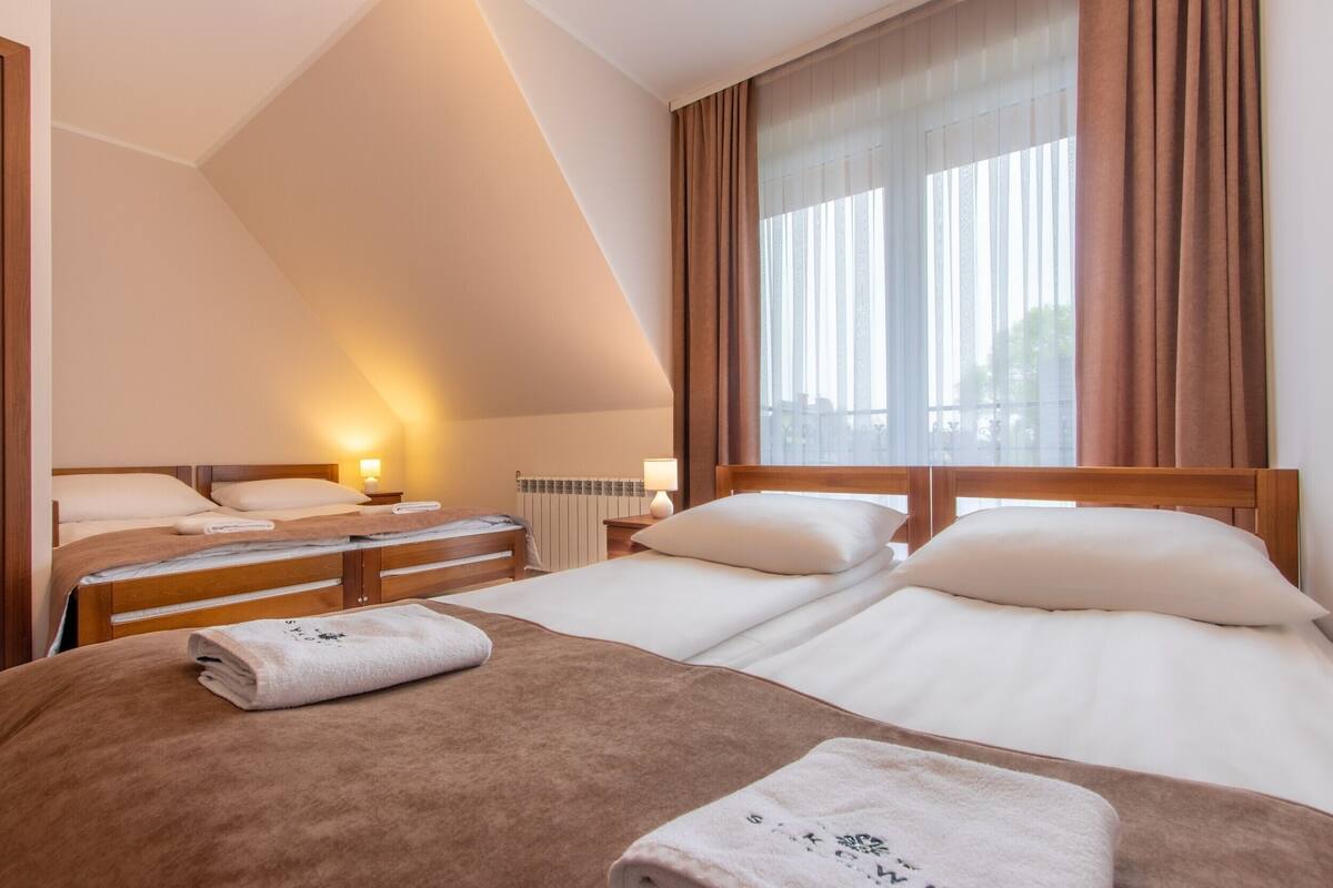 A hotel room with two beds, a large window with curtains, a radiator, and a nightstand with a lamp.