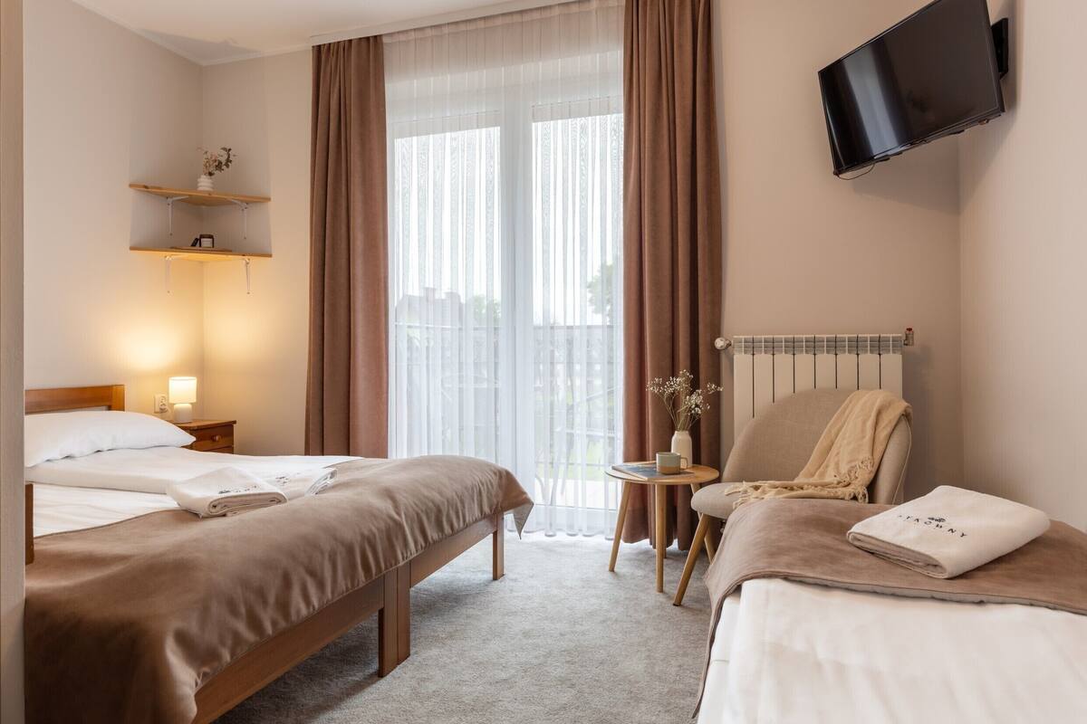 A hotel room with two beds, a TV mounted on the wall, a small table with a vase, and a chair.