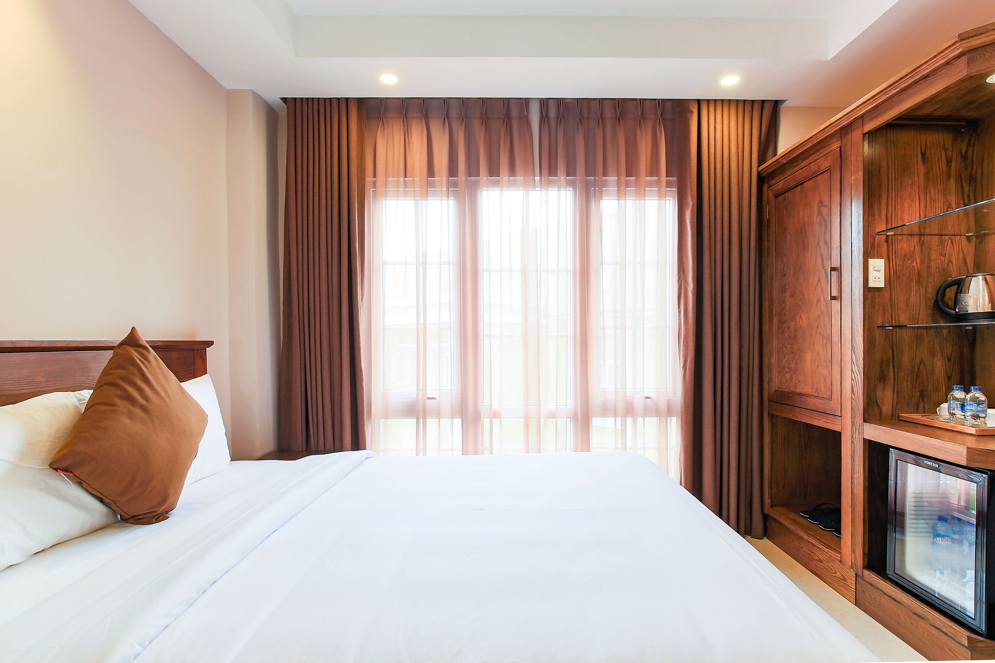A hotel room with a bed, curtains, a wooden wardrobe, and a minibar.