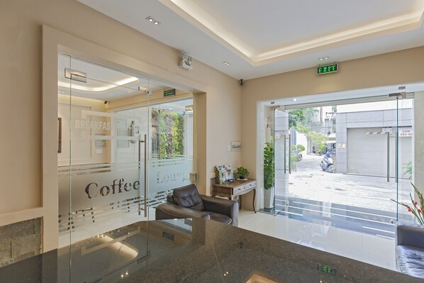 A modern lobby with a glass wall displaying a coffee shop sign, a leather sofa, a desk with a plant, and an exit sign. - Hoang Lan Hotel (Ho Chi Minh City)