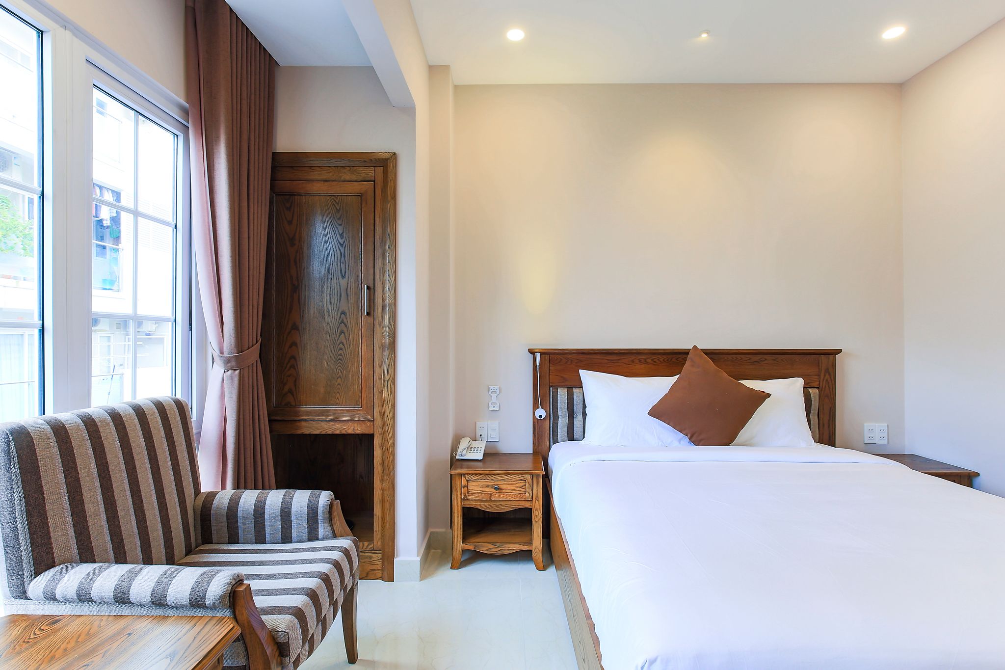 A hotel room with a bed, a wooden nightstand, a striped armchair, a wooden wardrobe, and a window with curtains.