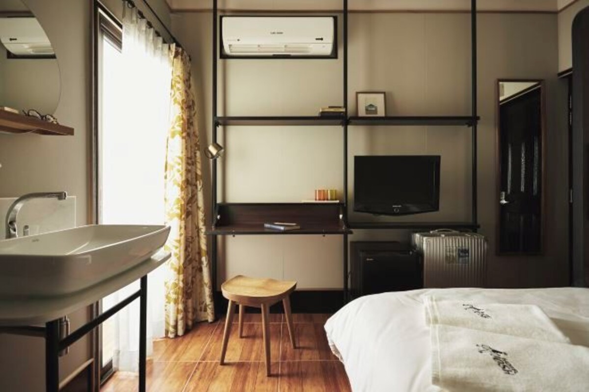 A hotel room with a bed, a bedside table, a TV, a desk with a chair, a window with curtains, and an air conditioning unit.