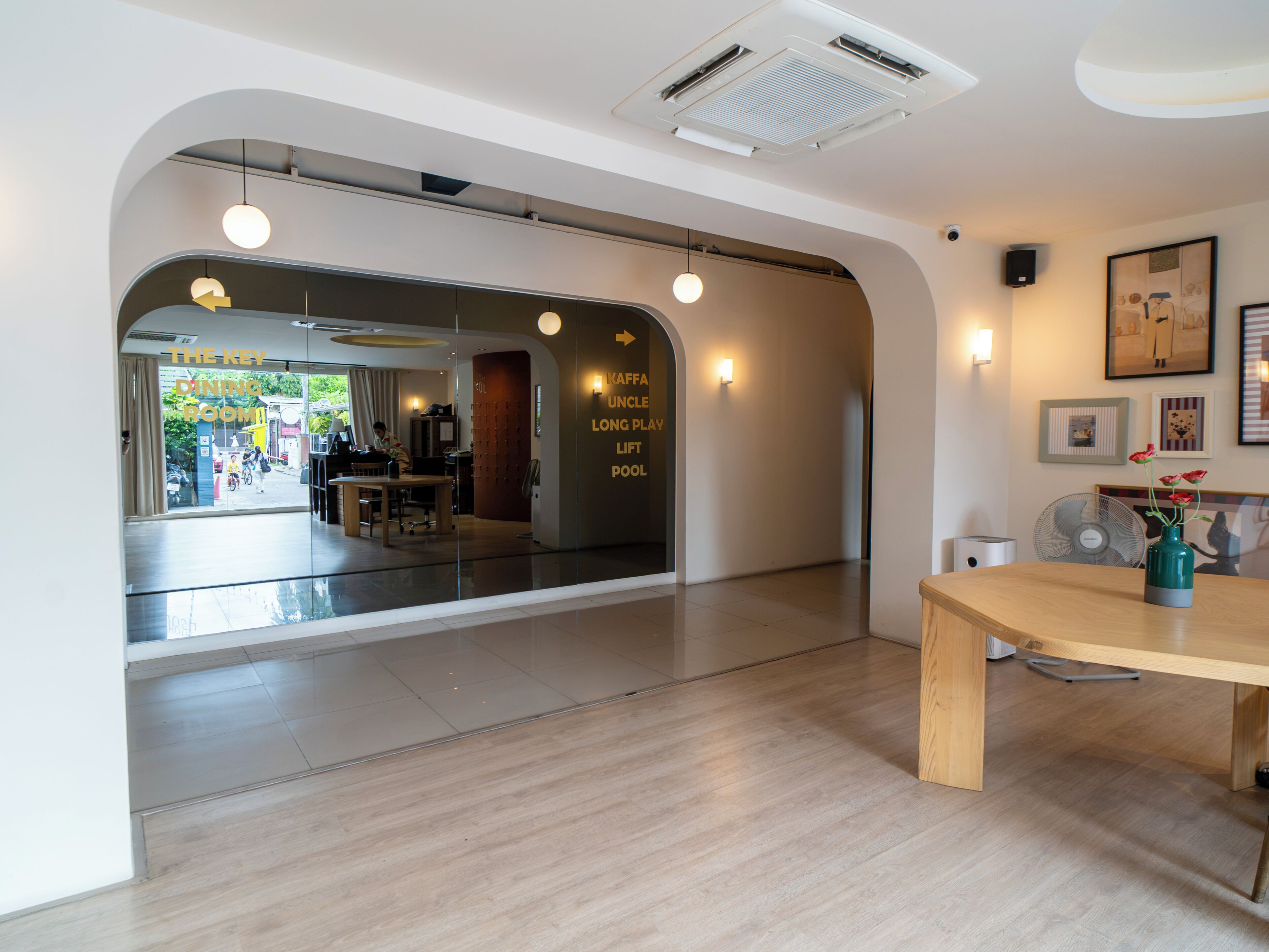 A hotel lobby with a dining area visible through a glass partition, a wooden reception desk, and a wall-mounted fan.