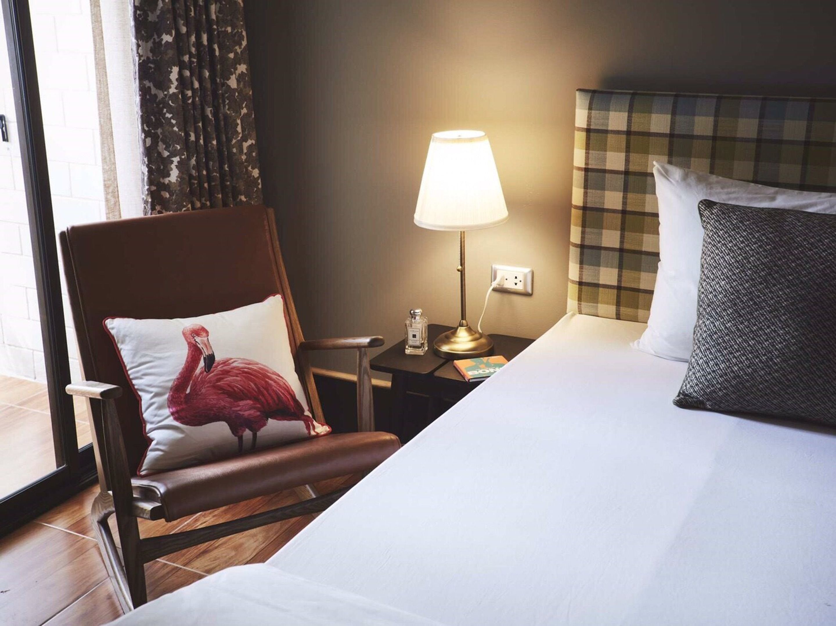 A hotel room with a bed, a nightstand with a lamp, a chair with a flamingo pillow, and a checkered headboard.