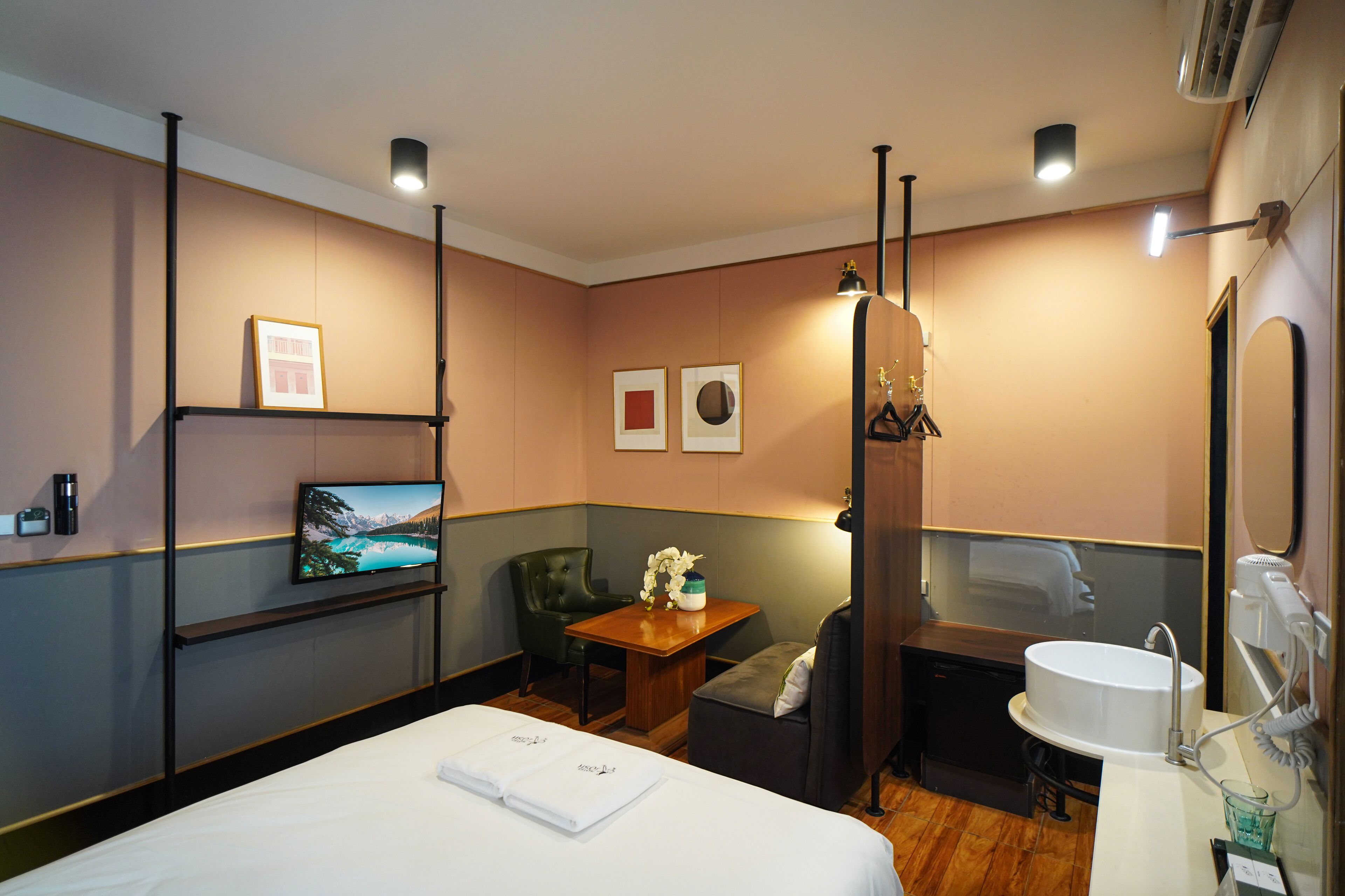 A hotel room with a bed, a desk, a chair, a small table with a vase, a mirror, and a sink.