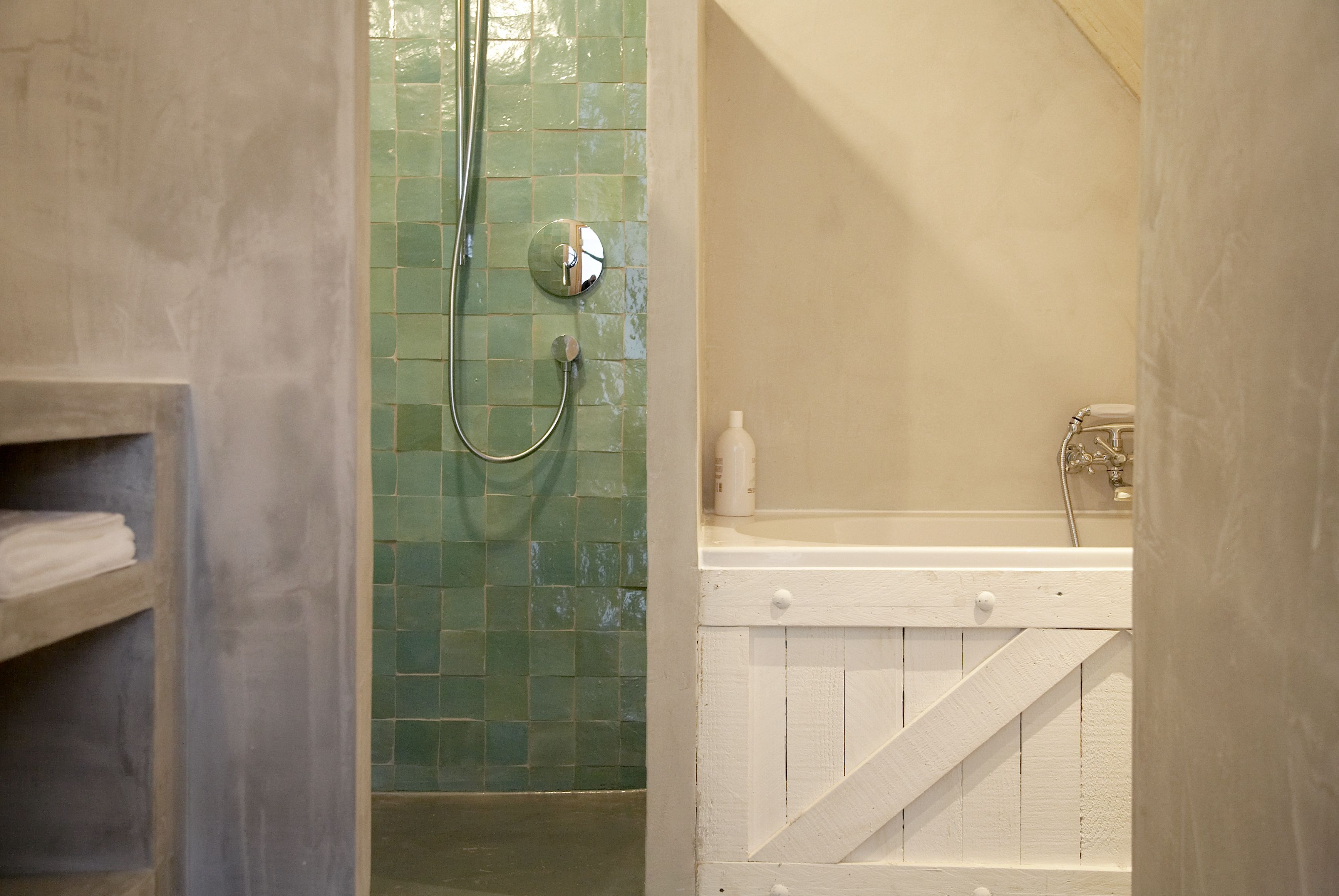 A bathroom with a shower area featuring green mosaic tiles, a white bathtub with a wooden accent, and a towel shelf.