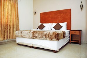 Una cama bien hecha con cabecera marrón, colcha blanca y dos almohadas marrones. A su derecha, una mesita de noche de madera con un libro encima.