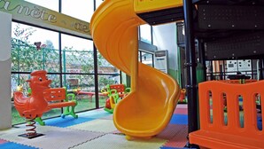Zona de juegos infantiles con un tobogán espiral amarillo, juegos coloridos y un ventanal grande que ofrece vistas al exterior.