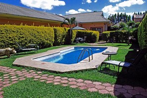 Una piscina rectangular con una escalera metálica, rodeada por un patio de ladrillo y césped verde. Hay sillas de descanso y una pequeña mesa cerca.