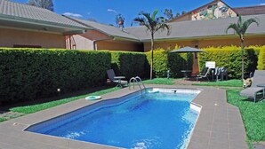 Una piscina rectangular con borde de azulejos, rodeada por un césped cuidado y un seto.