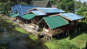 A riverside wooden house with a green roof, a dock, and a small deck with chairs. - Baan Suanaomsuk (Pak Tho)