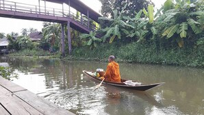 A monk in a boat on a calm river, with a bridge and lush greenery in the background. - Baan Suanaomsuk (Pak Tho)