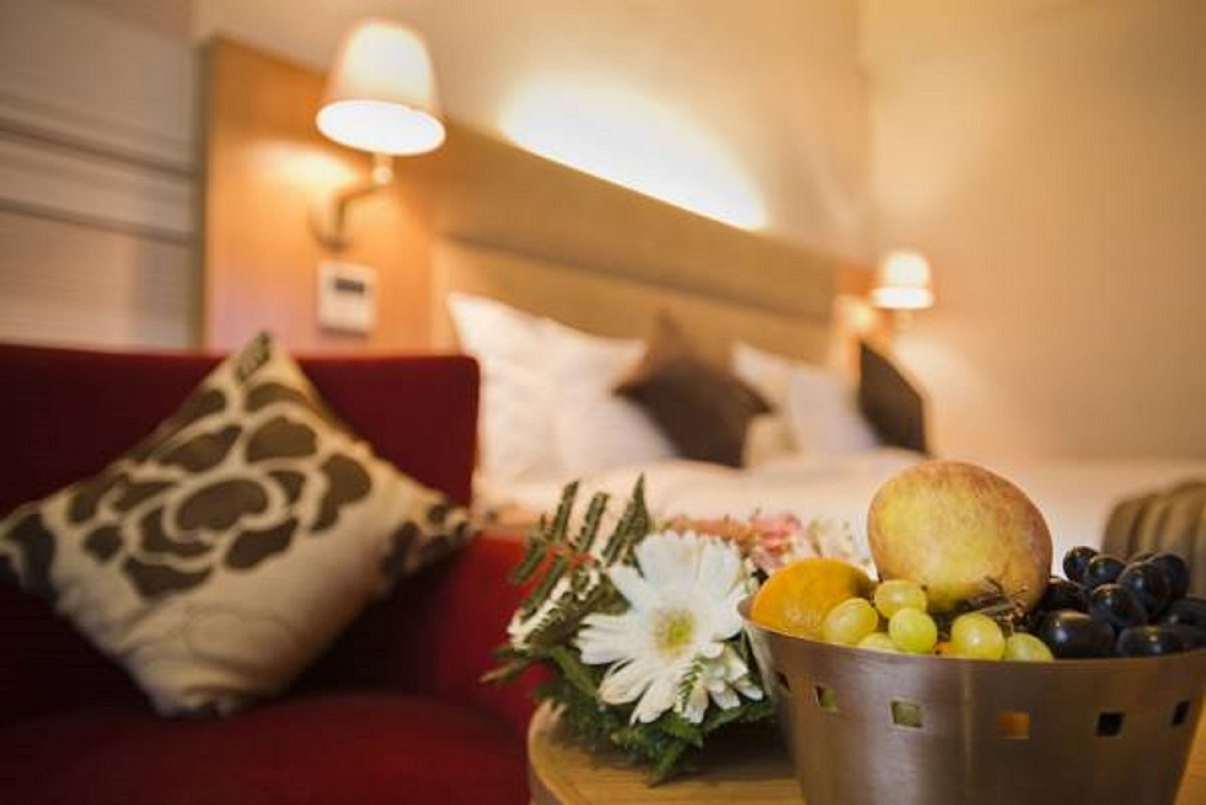 Habitación de hotel con cama, sofá rojo, almohada decorativa, frutera y lámpara de pared.