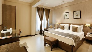 A hotel room with a large bed, a desk with a chair, a small table, and a bench.