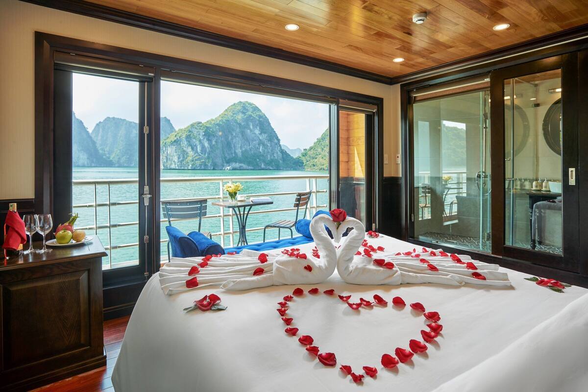 A hotel room with a bed decorated with swans and rose petals, a view of the sea and mountains, and a dining area with a table and chairs.