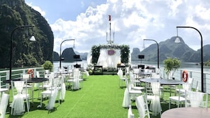 Montagem de casamento ao ar livre com tapete verde, cadeiras brancas e mesas dispostas para os convidados, com vista para um corpo de água e montanhas.