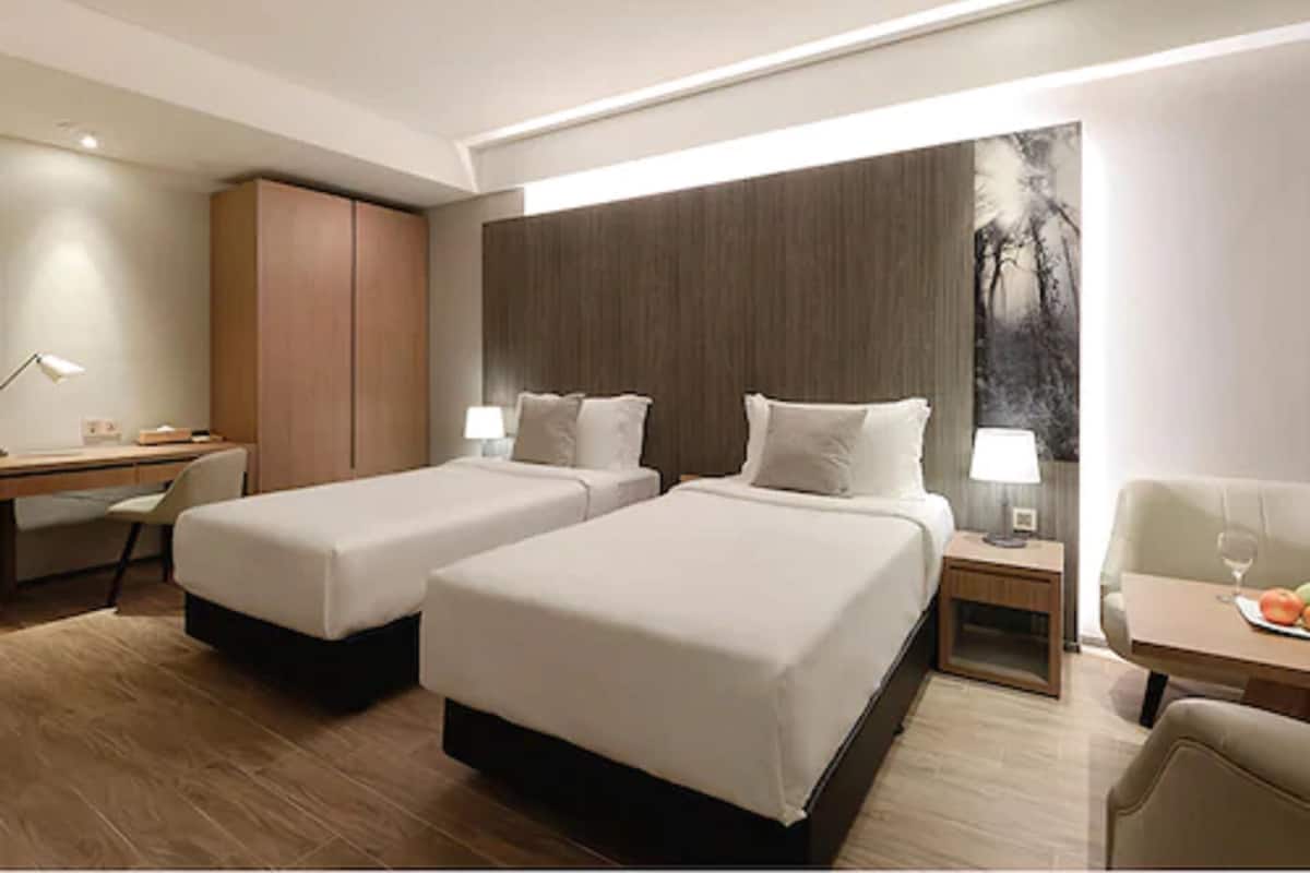 Habitación de hotel con dos camas, un escritorio, una silla, una mesita con un vaso y un cuadro en la pared.