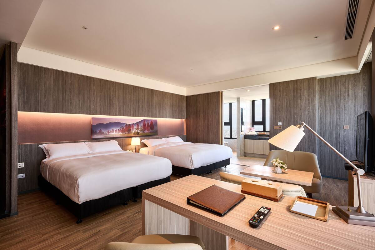 Habitación de hotel con dos camas, un escritorio con lámpara, un control remoto y una mesita.