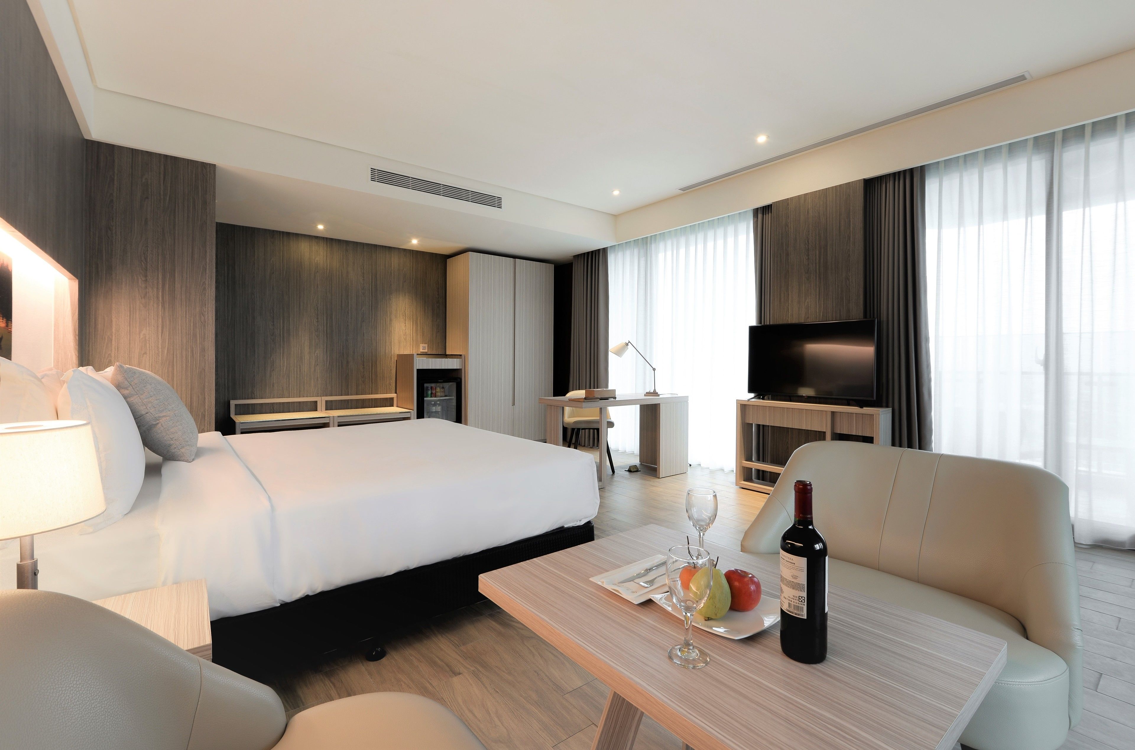 Een hotelkamer met een groot bed, een sofa, een eettafel met een fles wijn en fruit, en een televisie.
