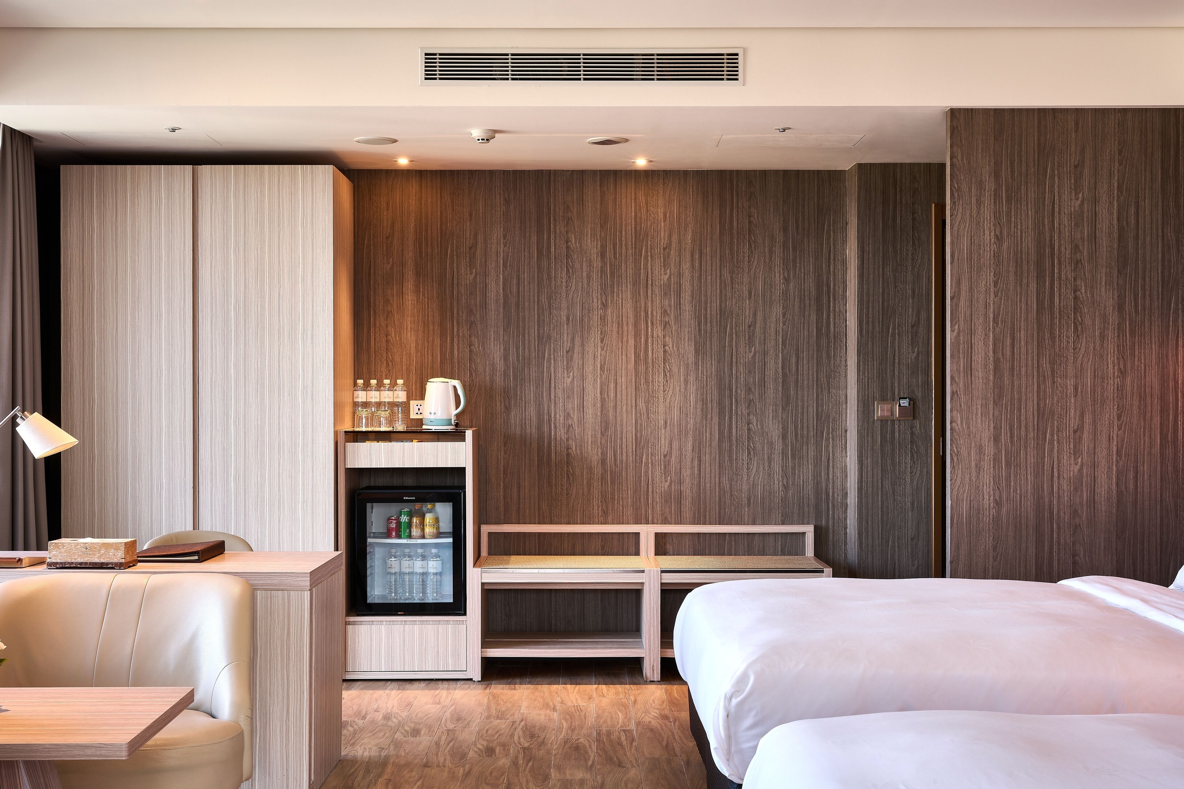 A hotel room with a bed, a desk with a lamp, a minibar, and a seating area.