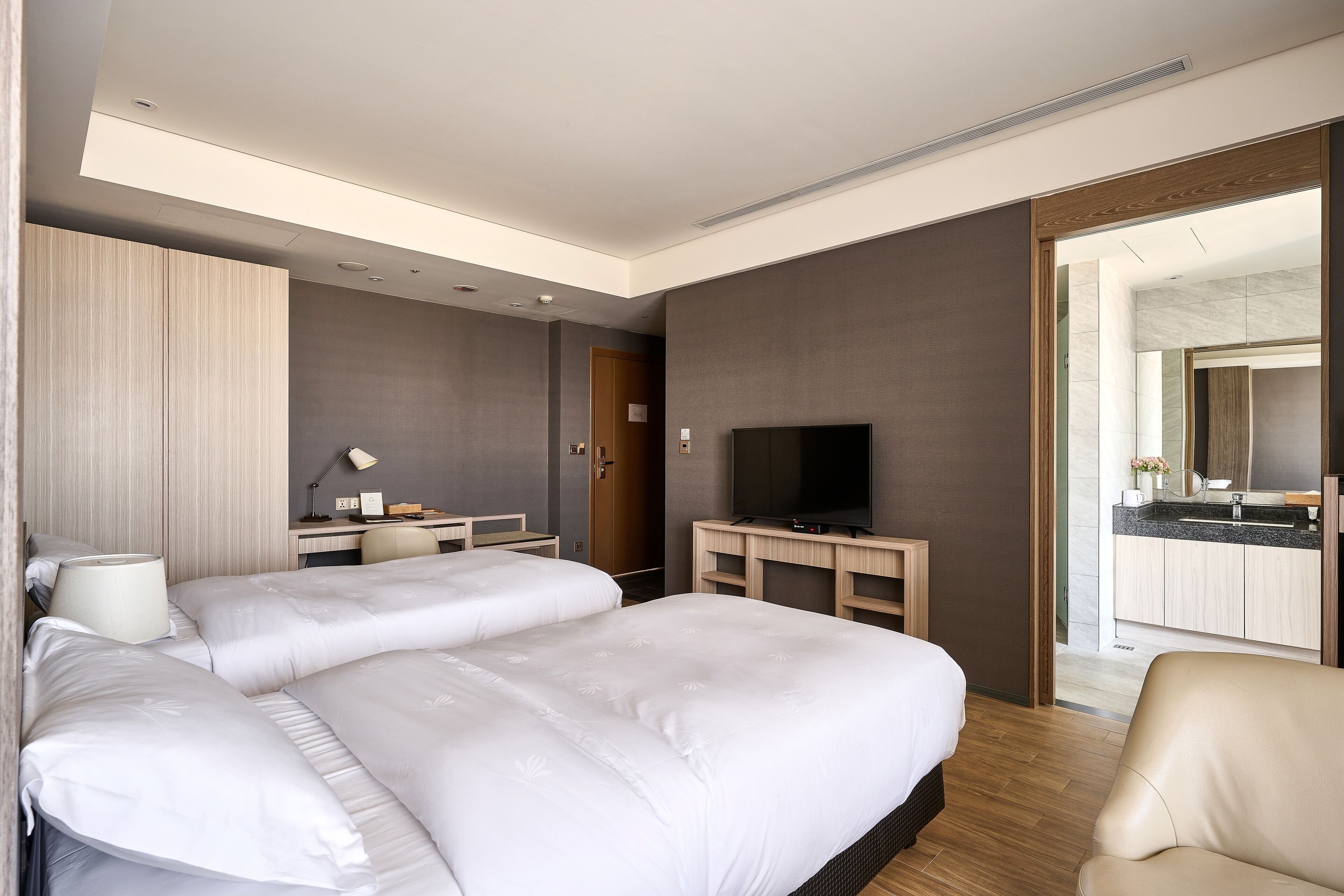 Habitación de hotel con dos camas, un escritorio con televisor de pantalla plana, una silla y un baño visible a través de una puerta abierta.