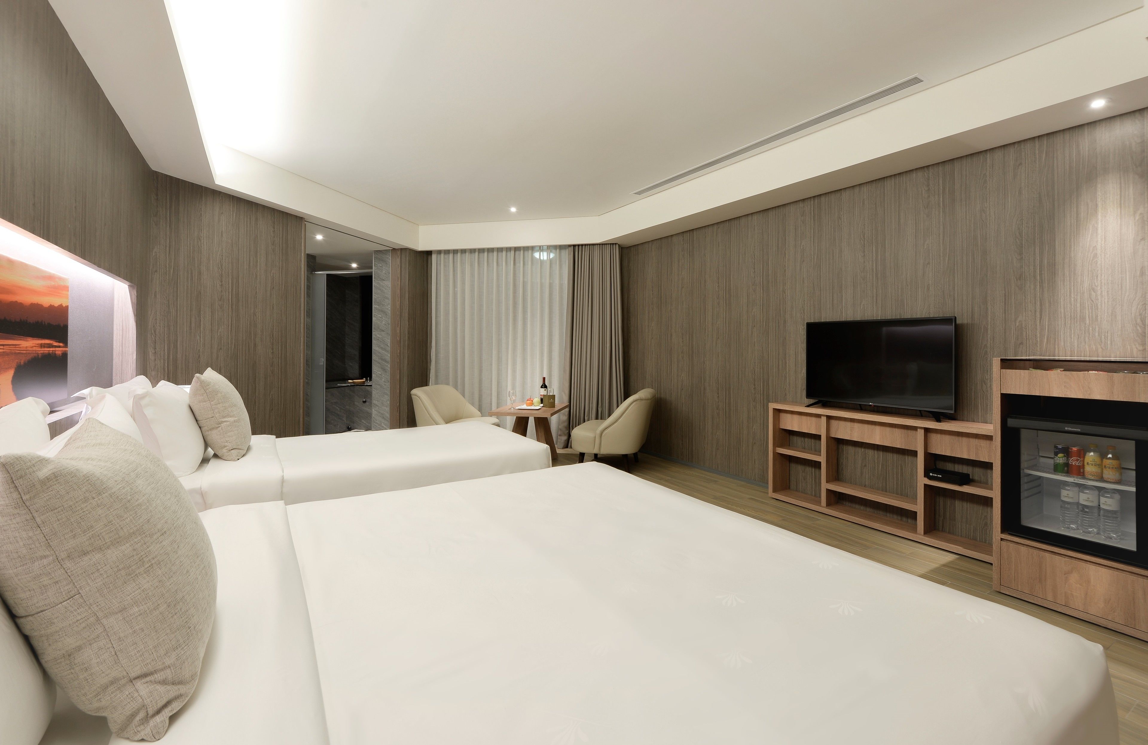 Ett modernt hotellrum med två sängar, en tv, en minibar och ett stort fönster med gardiner.