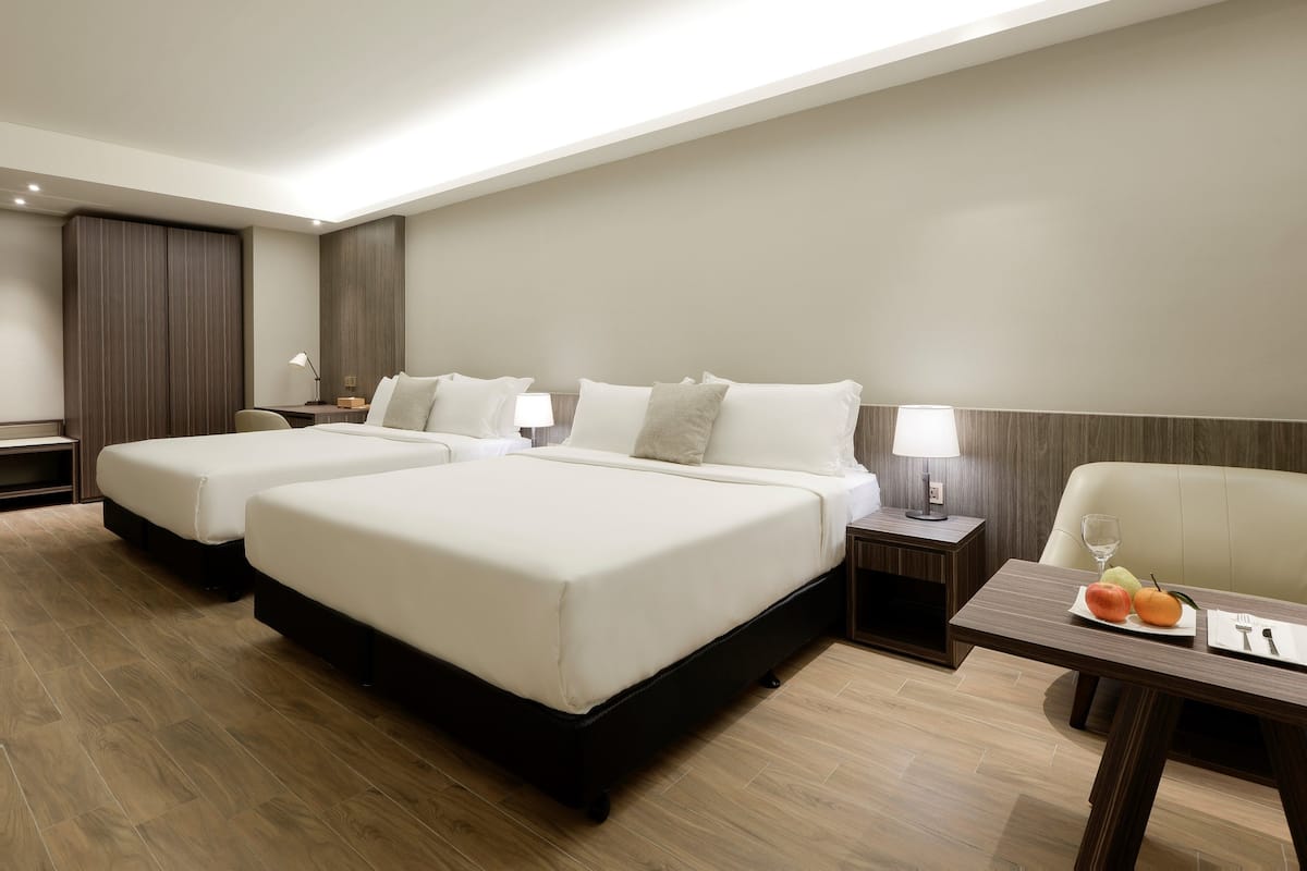 Habitación de hotel con dos camas, una mesa de comedor con fruta y un vaso, y una lámpara a cada lado de las camas.