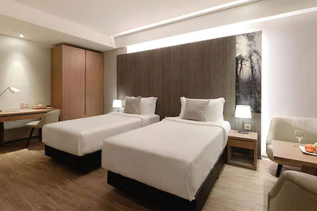 Habitación de hotel con dos camas, un escritorio, una silla, una mesita con un vaso y un cuadro en la pared.
