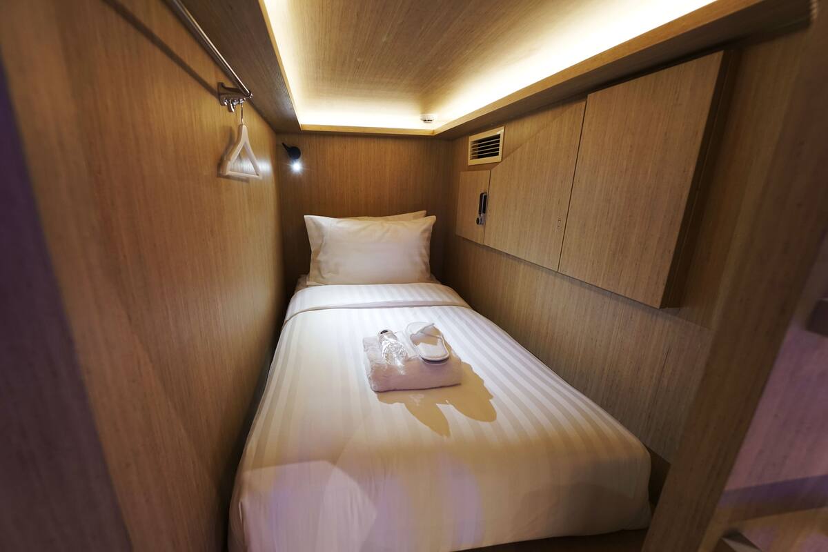 A compact, well-lit room with wooden walls, a single bed with white bedding, and a small bedside table.