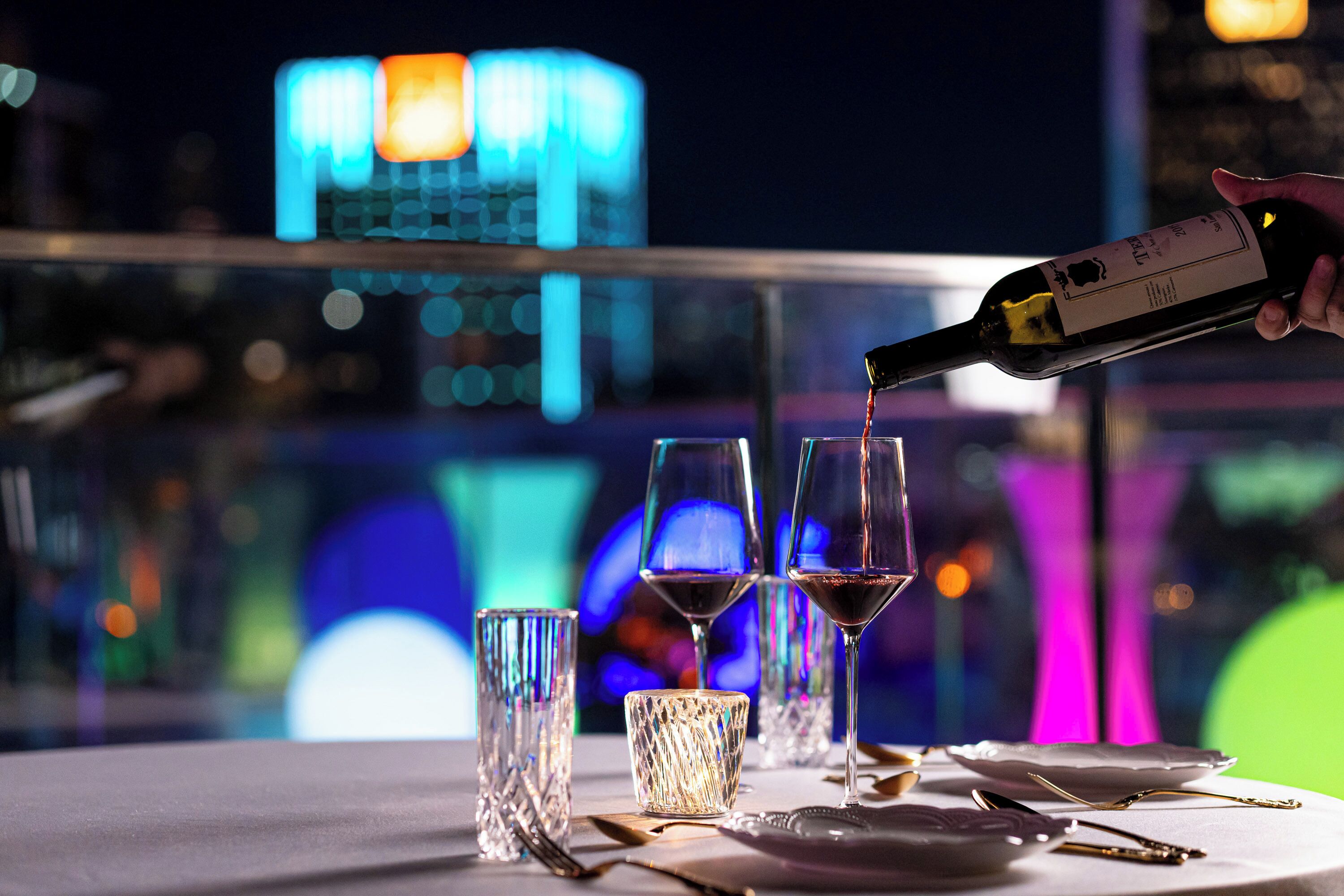 A bottle of wine being poured into glasses on a table with a cityscape view in the background.
