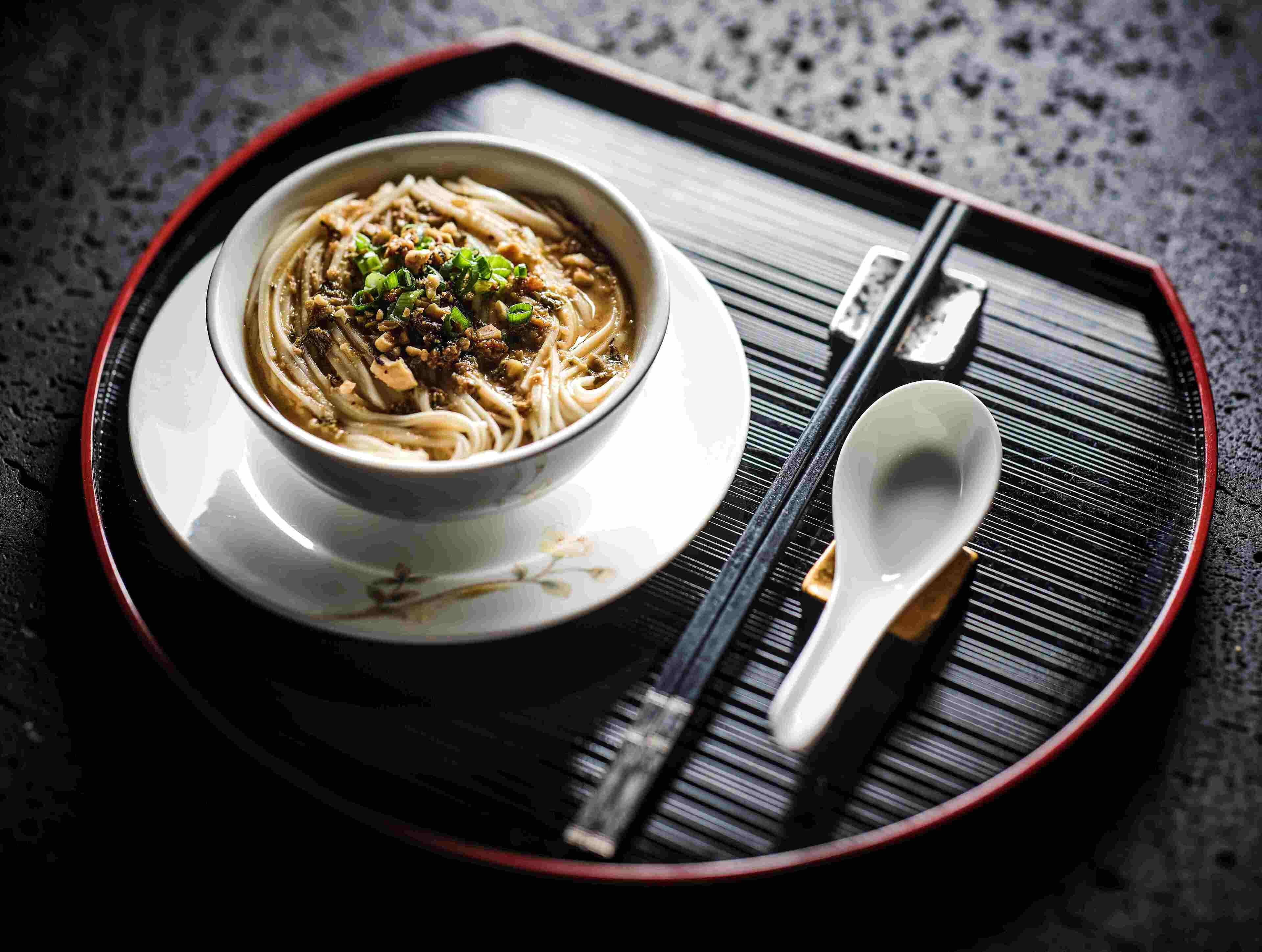 Eine Schüssel Ramen-Nudeln mit Belag auf einem Teller, zusammen mit Stäbchen und einem Löffel, platziert auf einer Tablett.