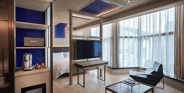 Quarto de hotel moderno com cama, televisão, máquina de café e área de estar perto de uma grande janela.