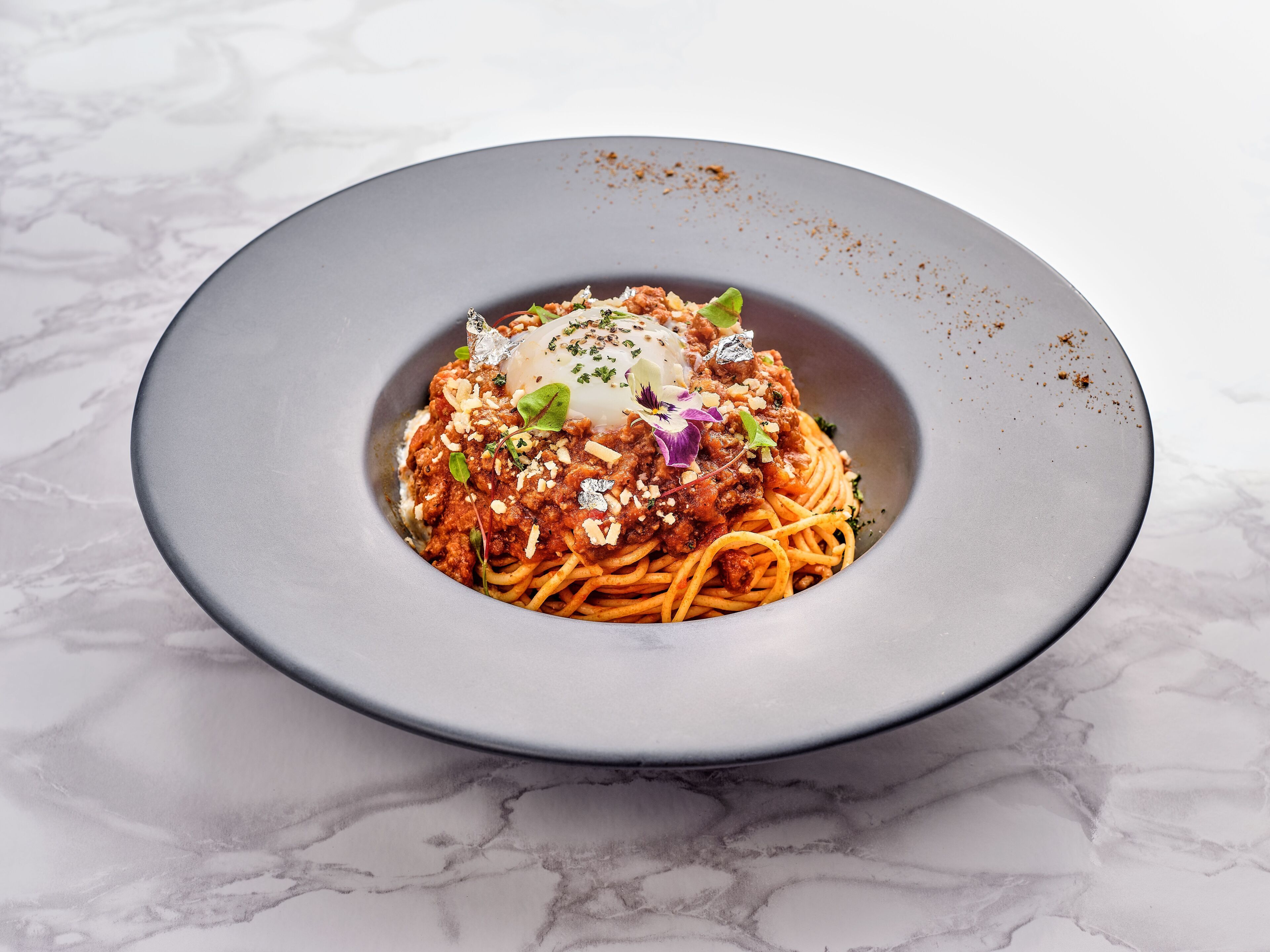 A plate of spaghetti with meat sauce, garnished with herbs and a poached egg, on a marble surface.
