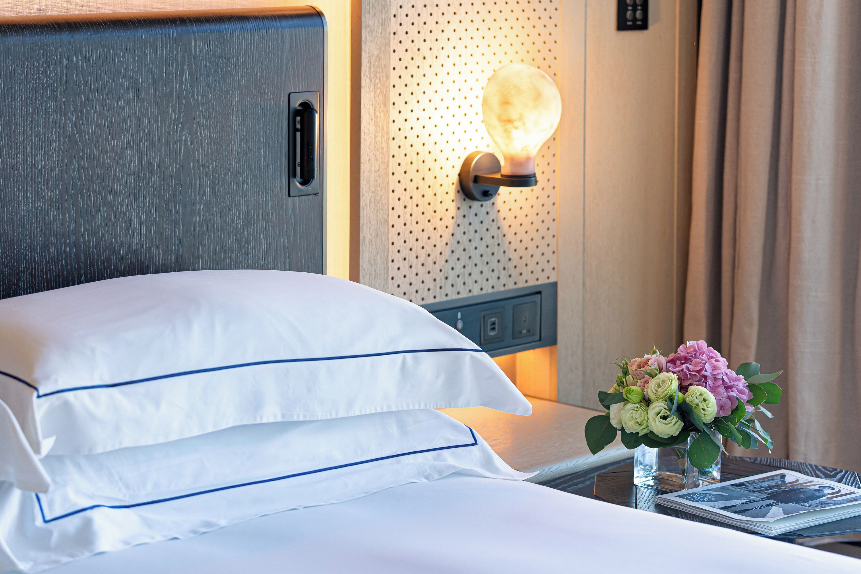 A neatly made bed with white linens and blue trim, a wall-mounted light fixture, a small vase with flowers, and a nightstand with a magazine.