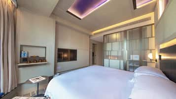 A modern hotel room with a large bed, a small table with a magazine, a TV, and a minibar.