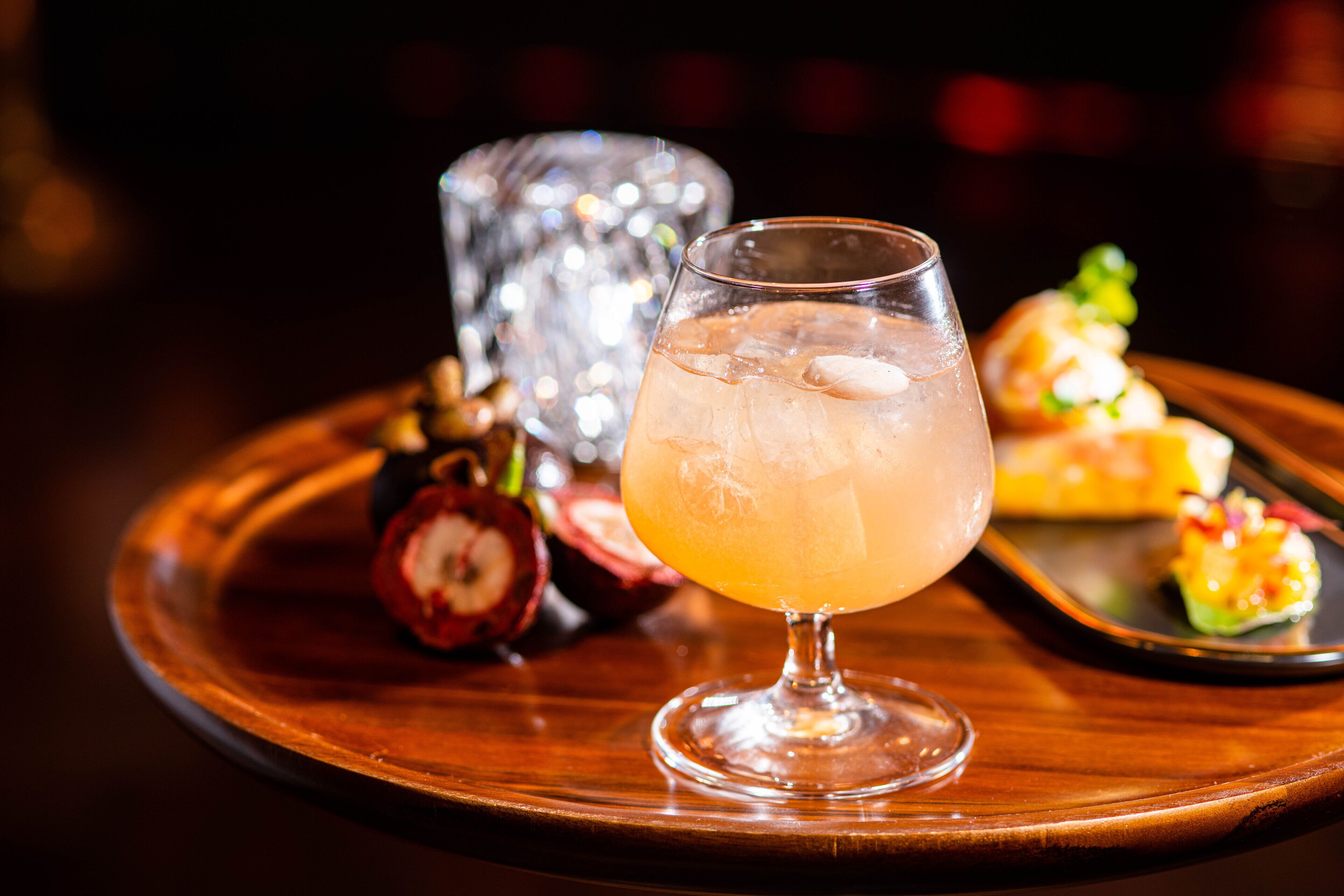 Un verre à cocktail rempli de glaçons, posé sur un plateau en bois, accompagné d’une tranche de fruit et d’un verre décoratif.