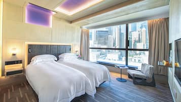 Habitación de hotel moderna con una cama grande, zona de estar y vistas a la ciudad a través de amplios ventanales.