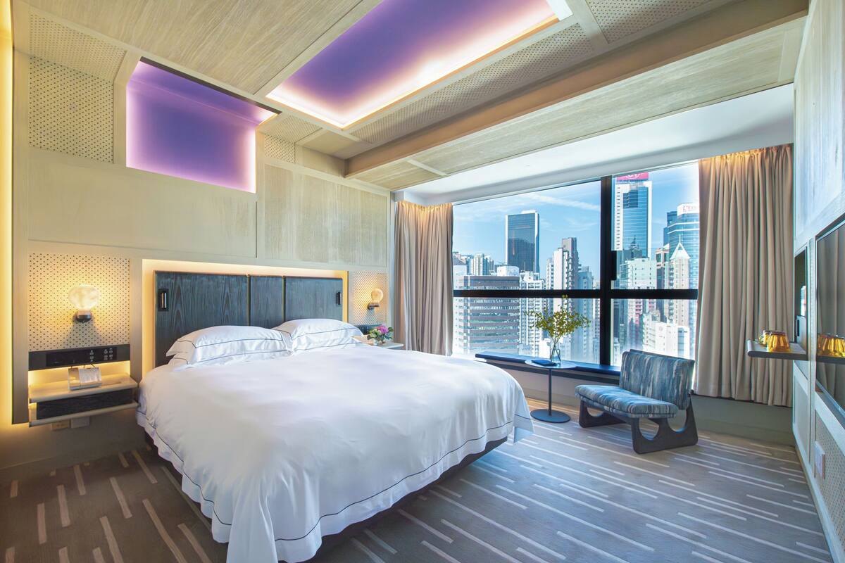 A modern hotel room with a large bed, a city view, and a seating area by the window.