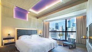 A modern hotel room with a large bed, a city view, and a seating area by the window.