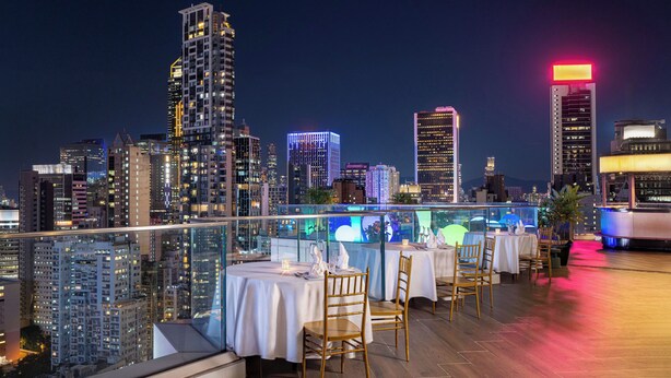 A rooftop dining area with tables and chairs, overlooking a cityscape with illuminated buildings.
